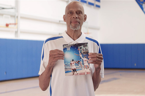 Kareem Abdul-Jabbar standing on basketball court holding a photo of him playing basketball - #WeLoveAHealthyLA